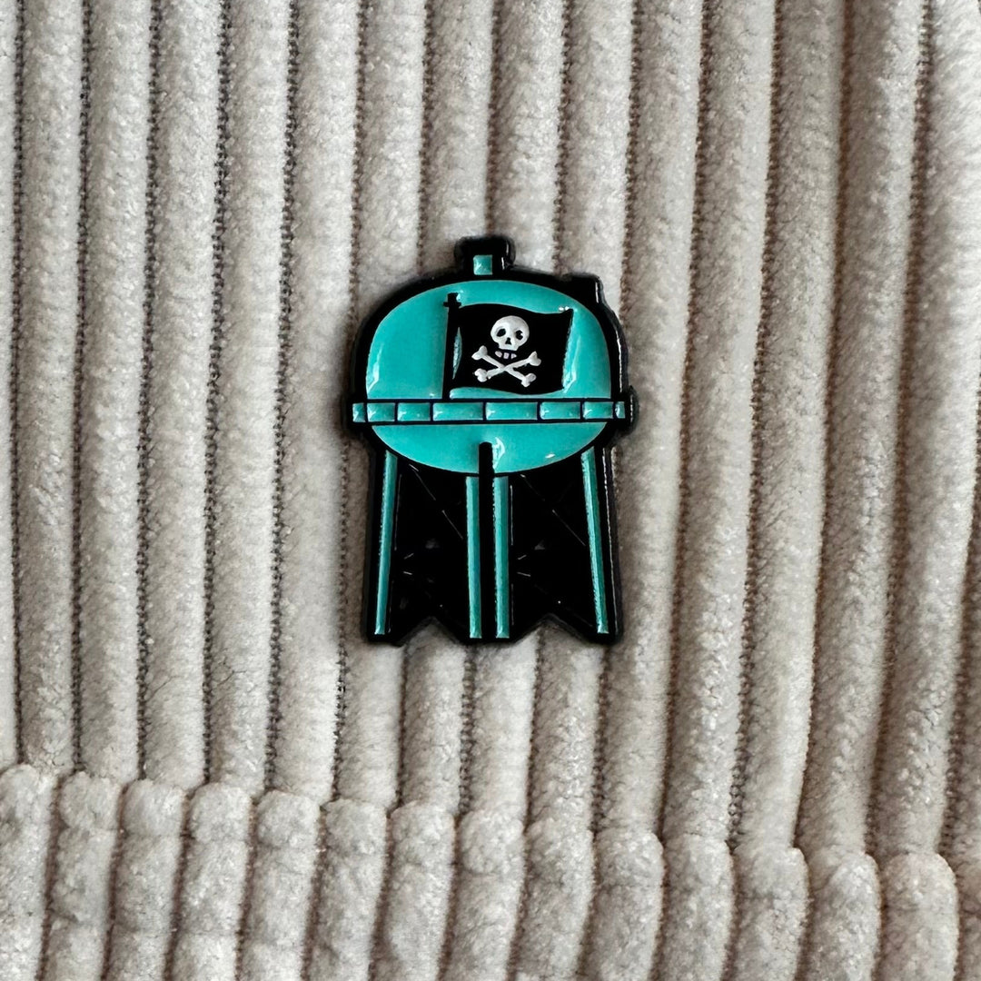 Teal and black enamel pin with skull design on a textured beige background