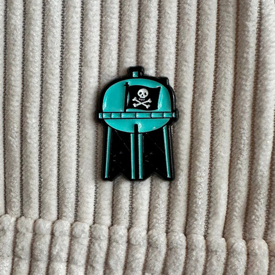 Teal and black enamel pin with skull design on a textured beige background