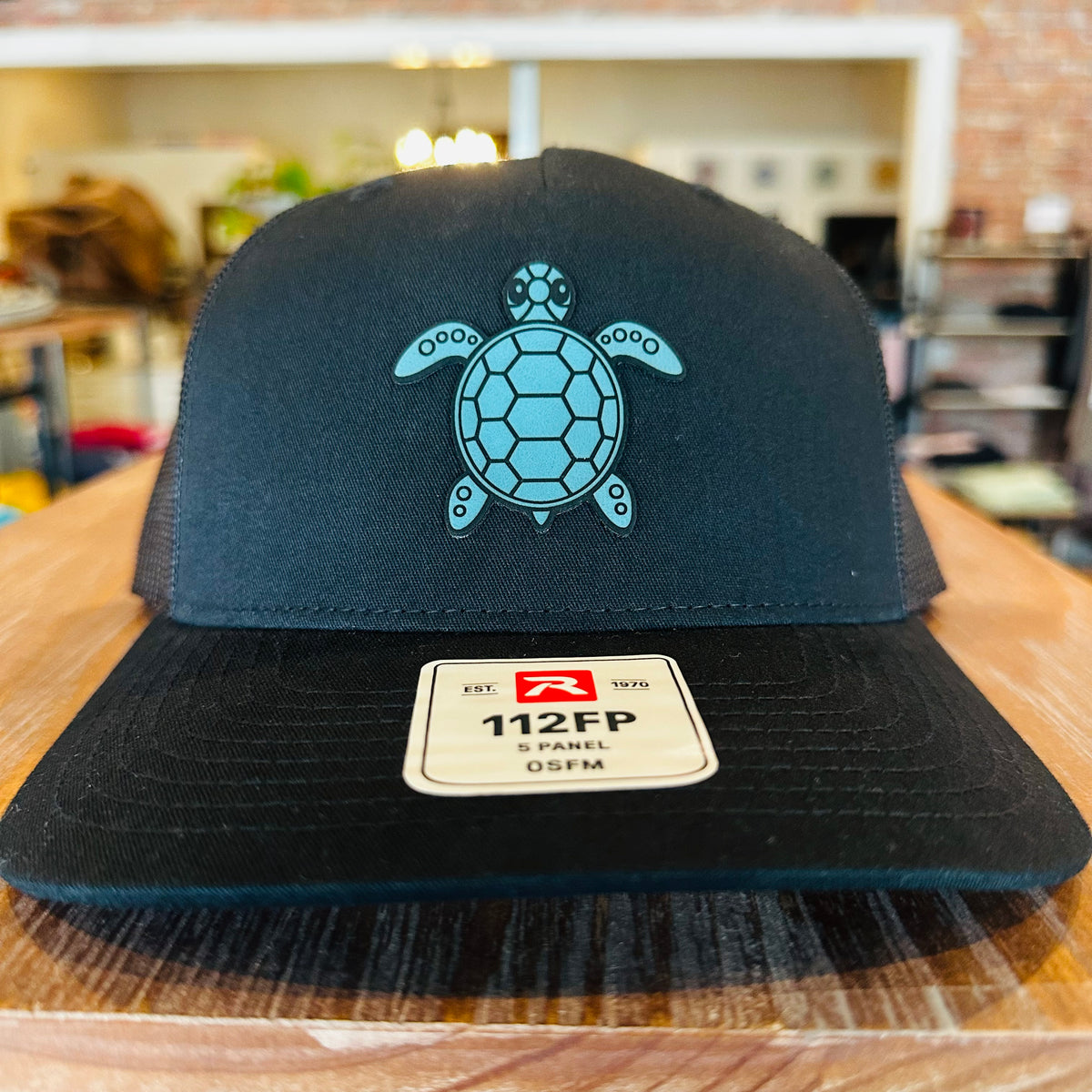 A black trucker-style cap with a turquoise sea turtle graphic on the front, resting on a wooden table. The brim features a white 112FP snapback label.