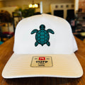 A white 5-panel trucker hat with a teal turtle graphic on the front, resting on a wooden table. The brim features a beige patch with the text '112FP 5 PANEL GSWM' and a red logo.