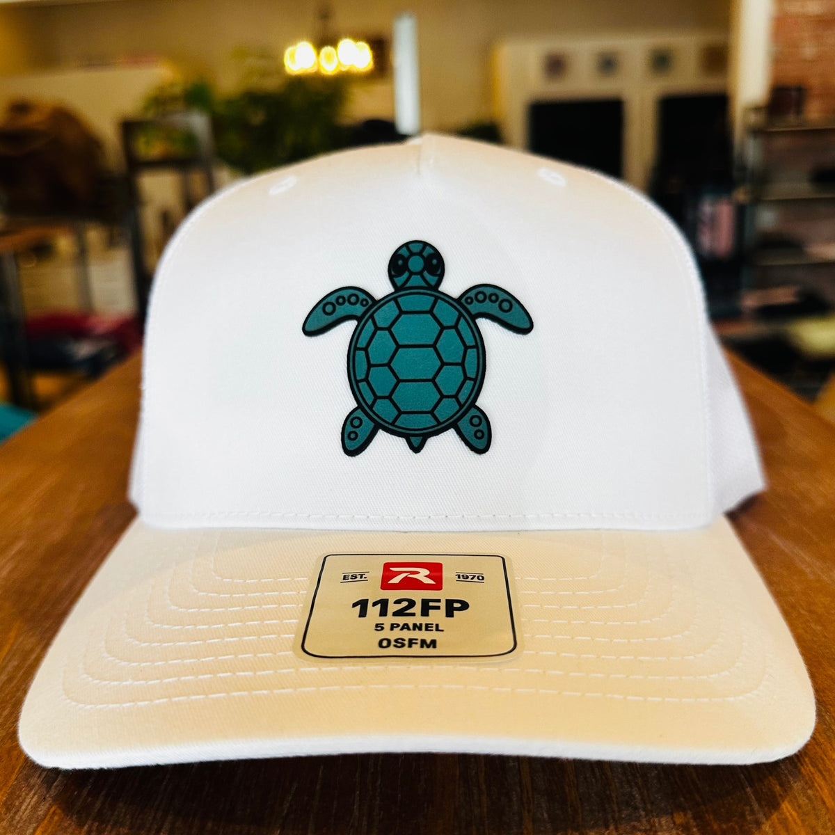 A white 5-panel trucker hat with a teal turtle graphic on the front, resting on a wooden table. The brim features a beige patch with the text '112FP 5 PANEL GSWM' and a red logo.