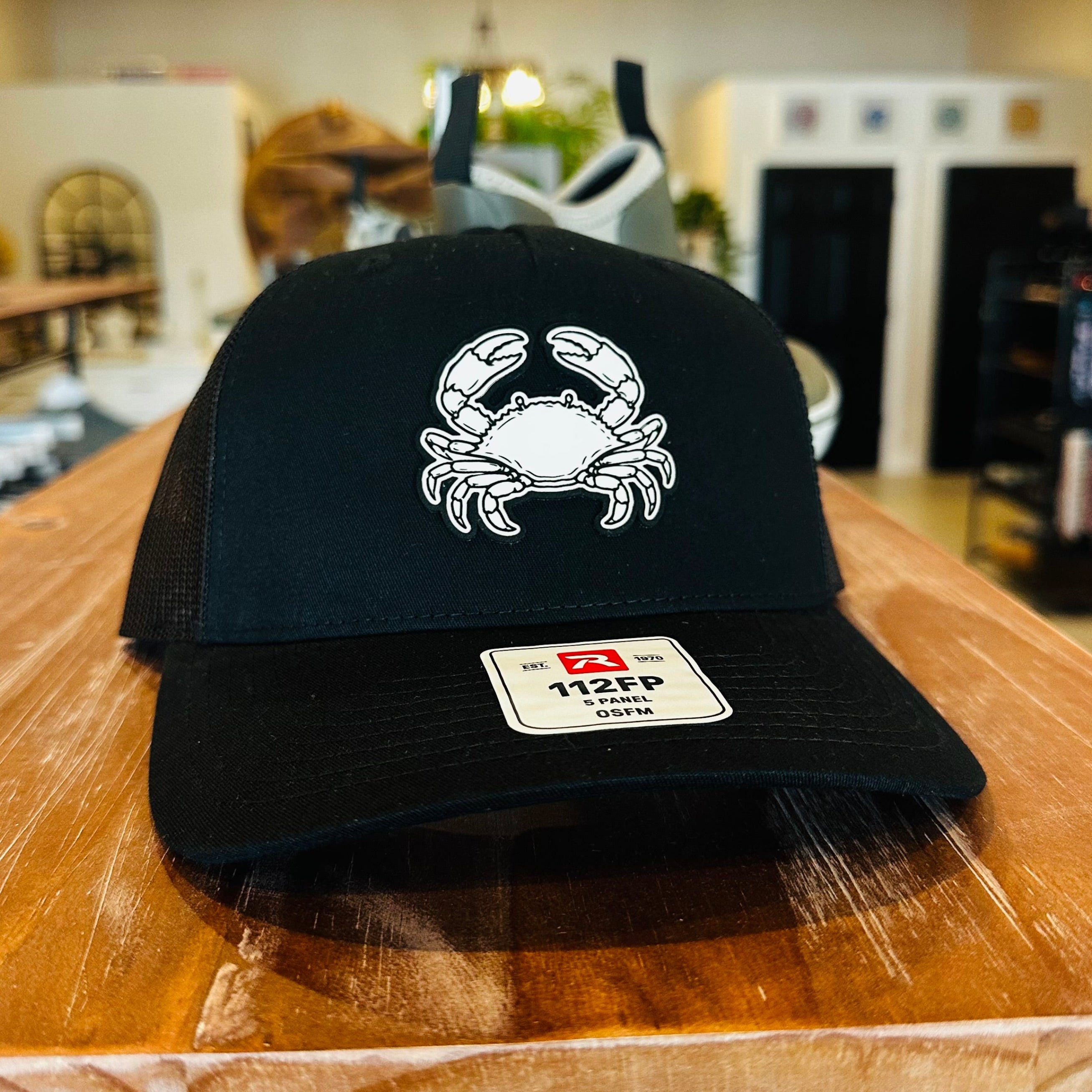 A black trucker-style baseball cap with a white embroidered crab graphic on the front, displayed on a wooden counter inside a store. The cap has a visible 112FP brand patch on the brim.