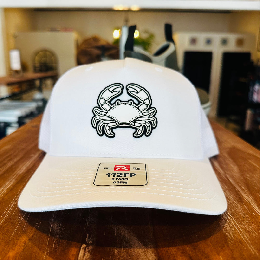 A white baseball cap with a black embroidered crab logo on the front, resting on a wooden table in a blurred indoor setting.