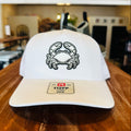 A white baseball cap with a black embroidered crab logo on the front, resting on a wooden table in a blurred indoor setting.