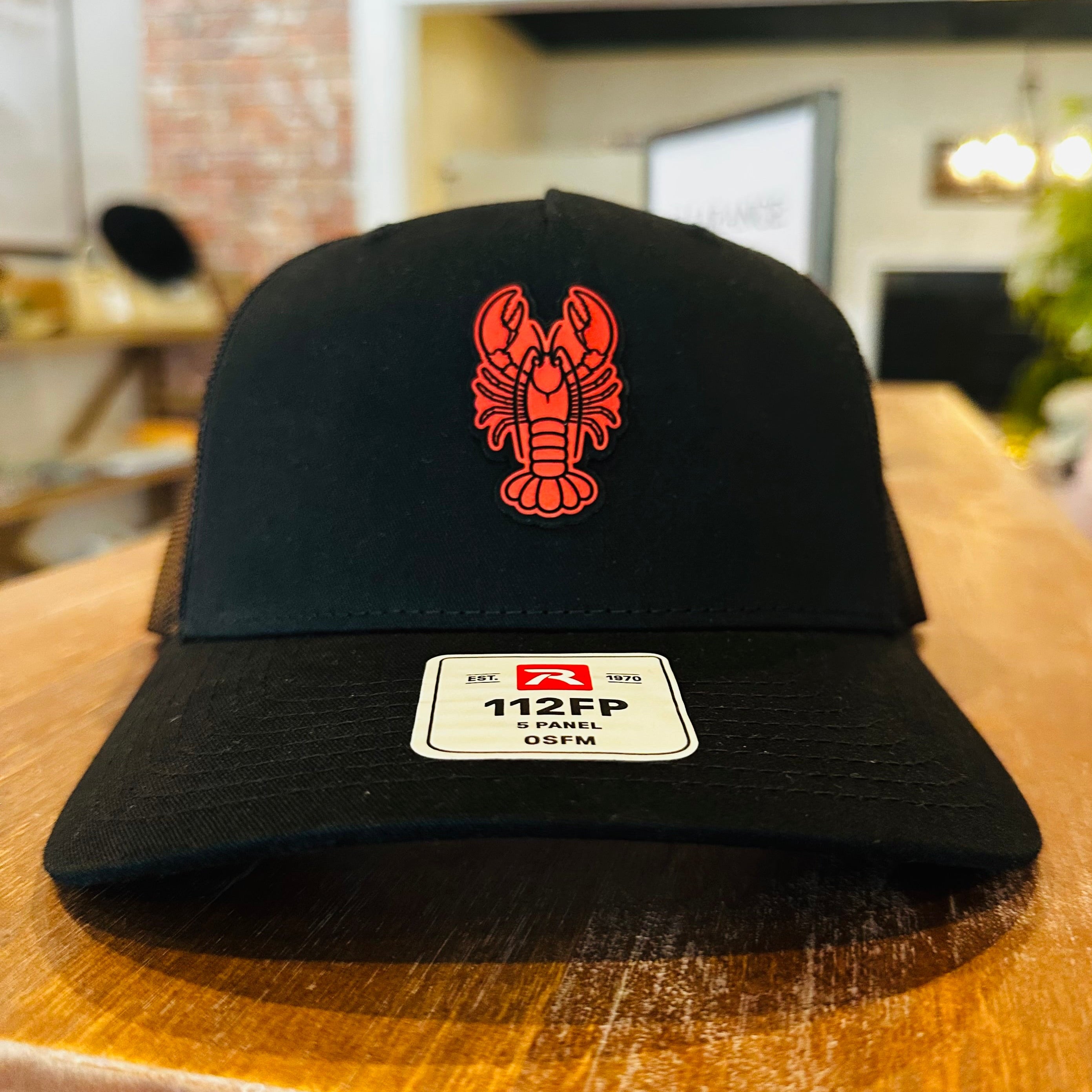 A black trucker-style cap with a red lobster logo on the front, resting on a wooden table in a store setting. The brim has a white size sticker reading 112FP.