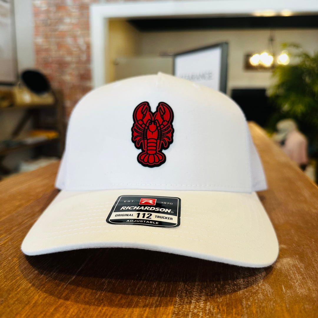A white Richardson 112 adjustable baseball cap with a red lobster logo on the front, resting on a wooden table in a blurred indoor setting.