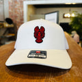 A white Richardson 112 adjustable baseball cap with a red lobster logo on the front, resting on a wooden table in a blurred indoor setting.